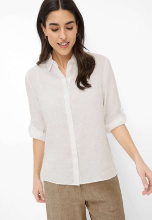 Donna con indosso una camicia bianca a maniche lunghe con maniche arrotolate e pantaloni marrone chiaro, sorridente e con lo sguardo rivolto verso il basso.