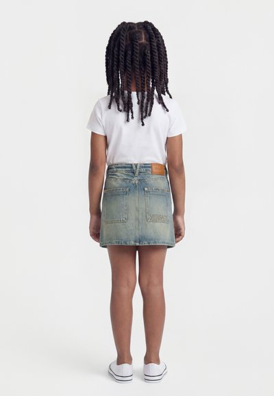 Παιδί με πλεκτά μαλλιά, που στέκεται γυρισμένο με την πλάτη, φοράει λευκό μπλουζάκι, τζιν φούστα και λευκά αθλητικά παπούτσια, πάνω σε απλό φόντο.