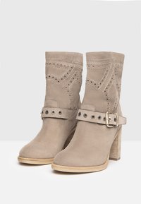 Beige Wildleder-Stiefeletten mit durchbrochenen geometrischen Mustern, einem dekorativen Riemen mit Metallösen und Schnalle sowie einem gestuften Blockabsatz.