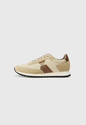 Beige sneakers with brown accents, suede and textile materials, round toe, flat black rubber sole, and lace-up design with logo detail.