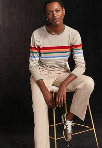 Pull beige avec des rayures horizontales multicolores, associé à un pantalon beige clair et des baskets marron. Cheveux courts, assis sur un tabouret.