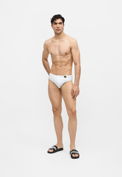 Νέος άνδρας με λευκά μαγιό briefs και μαύρα slides Moschino που στέκεται μπροστά σε ένα απλό φωτεινό φόντο.