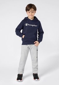 Tengerészkék kapucnis pulóver fehér logóval, szürke logónyomatos melegítőnadrággal párosítva, fekete sportcipő narancssárga részletekkel.