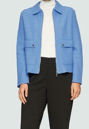 Femme portant une veste zippée bleu clair avec poches à rabat sur un col roulé blanc et un pantalon noir, debout devant un fond uni.