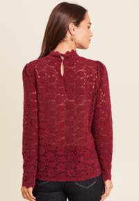 Burgundy Spitze Bluse mit langen Ärmeln, gewelltem Ausschnitt und Schlüsselaugen-Detail hinten, das ein strukturiertes florales Muster aufweist.