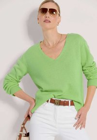 Grüner V-Ausschnitt-Pullover mit gerippten Bündchen und Saum, kombiniert mit weißen Jeans und einem braunen Gürtel mit goldener Schnalle, ergänzt durch eine Sonnenbrille.