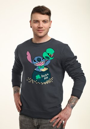 Homme portant un sweat-shirt sombre avec le personnage de dessin animé Stitch tenant un chapeau vert et sortant d'une marmite étiquetée "Lucky Me", avec des céréales éparpillées.
