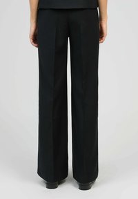 Personne portant un pantalon noir à jambes larges et des chaussures noires, vu de dos sur un fond gris uni.