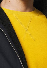 Gul tröja med en strukturerad yta har en framträdande korsstygnsdetalj. Den är lager-på-lager under en mörk, mjuk fleecejacka.