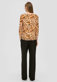 Multikleurige blouse met een abstract patroon in tonen van oranje, zwart en crème, met een ronde hals en ¾ mouwen. Gecombineerd met zwarte broek.