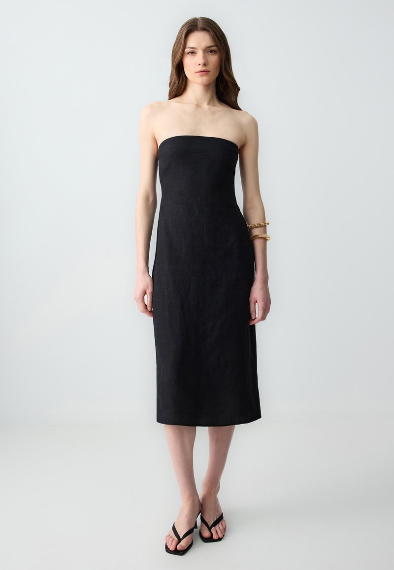Schwarzes, trägerloses Leinenkleid mit eng anliegendem Oberteil, knielangem Saum und glatter Textur. Kombiniert mit schwarzen Flip-Flops und einem goldenen Armreif.