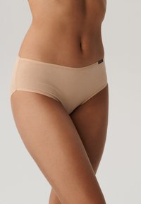 Beżowe, bezszwowe majtki o średnim stanie, gładkiej teksturze i minimalnym logo na pasie, zapewniające dopasowany i komfortowy design.
