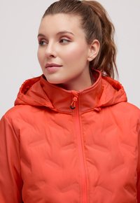 Oranžová zateplená bunda se zapínáním na zip, kapucí a texturovanou látkou. Mezi vlastnosti patří hladký povrch a prošité detaily.