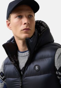 Mladý muž nosící námořnickou čepici, námořnickou zateplenou vestu s kruhovým logem a šedý svetr s modrými vzory, hledící mírně nahoru a doprava.