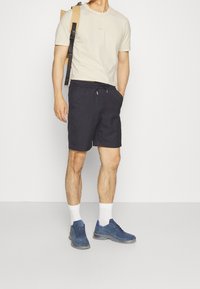 Beige bavlněné tričko s kulatým výstřihem, spárované s námořnickými šortkami s elastickým pasem. Modré tenisky a bílé ponožky dotvářejí vzhled.
