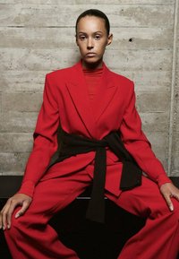 Woman with slicked-back hair wearing a red suit and black sweater tied around her waist, seated against a concrete wall.