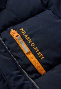 Sininen puhvijakku, jossa on oranssi vetoketju. Mukana on tasku, jossa on harmaa vetoketjun nyöri ja keltainen ompelu, jossa lukee "Polarn O. Pyret."
