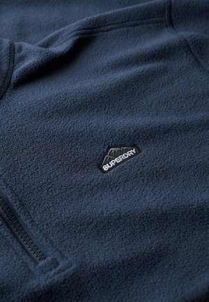 Pull en polaire bleu marine avec une poche zippée et un petit logo noir "Superdry" sur la poitrine. Tissu doux et texturé.