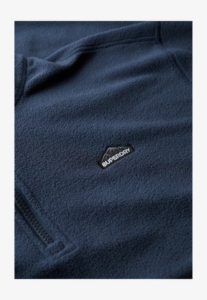 Navy blue fleece pullover featuring a zippered pocket and a small black "Superdry" logo on the chest. Soft, textured fabric.