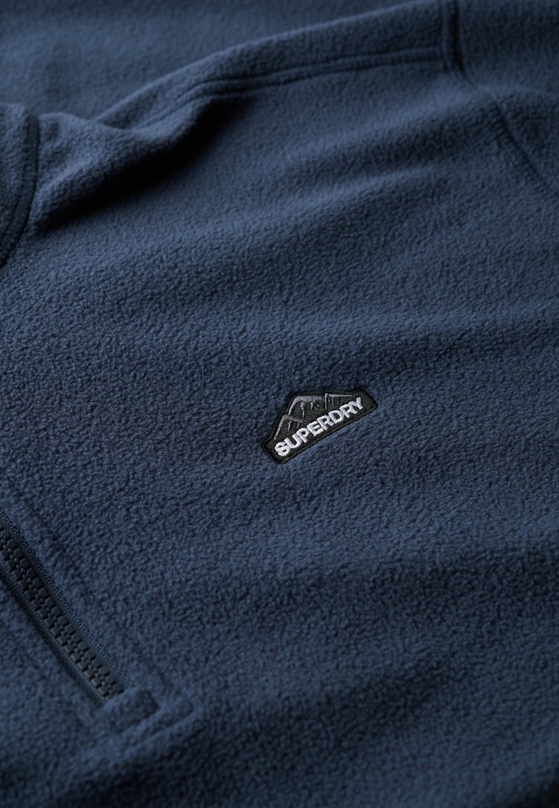 Navyblauwe fleece pullover met een ritszak en een klein zwart "Superdry" logo op de borst. Zacht, gestructureerd fabric.
