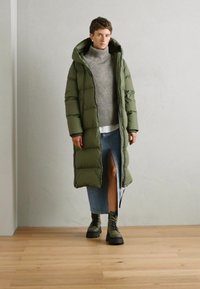 Abrigo tipo puffer largo de color verde oliva con capucha, suéter de punto gris y falda de mezclilla con una abertura lateral, combinados con botas de cordones de color verde oliva.