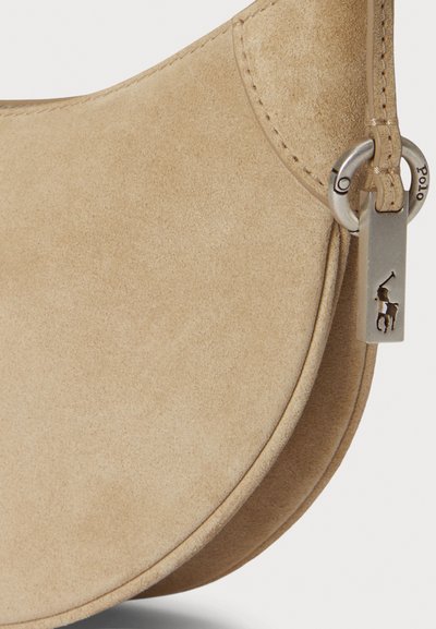 Beżowa zamszowa torebka na ramię o zaokrąglonym kształcie. Wyposażona w srebrne okucia, zawieszkę z logo oraz detale w postaci przeszyć dodających faktury.