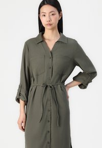 Vestido camisa verde-oliva com gola, mangas longas, abertura frontal com botões, dois bolsos no peito e um cinto na cintura.