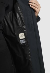 Sort yderjakke med en glat, skinnende inderside. Har et mærke med tekst og et iøjnefaldende logo. Fremstillet af slidstærkt stof.