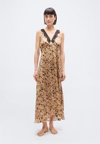 Γυναίκα με κοντά σκούρα μαλλιά, που φοράει αμάνικο σατέν φόρεμα με λεοπάρ σχέδιο και μαύρη δαντέλα στη φόδρα, στέκεται ξυπόλητη μπροστά σε ένα απλό φόντο.