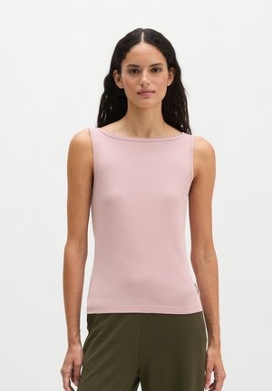 Vrouw met lang donker haar, gekleed in een mouwloos lichtroze top en olijfgroene broek, staand tegen een effen witte achtergrond.