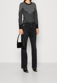 Schwarzer, gemusterter Pullover mit gestreiftem und geometrischem Design, kombiniert mit dunklen Jeans und spitzen, ledernen Absätzen. Die Accessoires umfassen eine kleine schwarze Tasche.