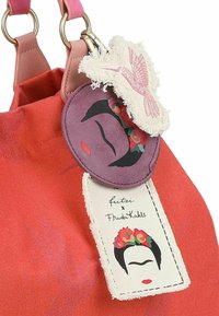 Sac en toile rouge avec des poignées roses, des anneaux en or et des patchs décoratifs représentant des motifs floraux et un visage stylisé.