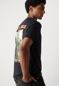 Schwarzes Baumwoll-T-Shirt mit einem übergroßen Grafikdesign von Kakteen auf dem Rücken und orangefarbener Schrift "NICHT ANFASSEN" darüber. Kurze Ärmel, lockere Passform.