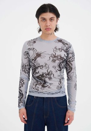Giovane con capelli castani di media lunghezza indossa un top grigio aderente a maniche lunghe con disegni neri di draghi e ragnatele e pantaloni larghi blu scuro.