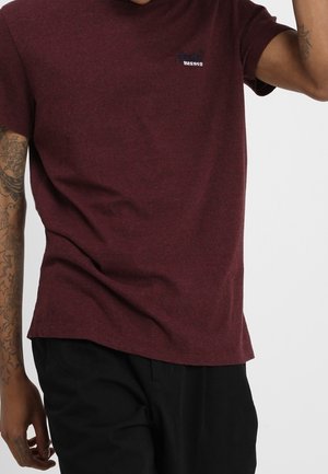 Homme portant un t-shirt à manches courtes bordeaux avec un petit logo sur la poitrine et un pantalon noir, montrant des tatouages sur le bras gauche.