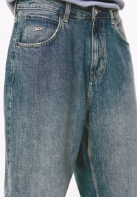 Primo piano di jeans in denim blu sbiadito con una spilla metallica del marchio sulla tasca anteriore e dettagli di cucitura visibili.