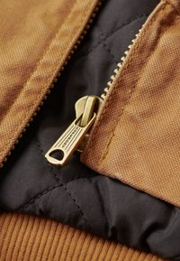 Tejido marrón texturizado con una cremallera dorada sobre un fondo acolchado negro. Presenta materiales contrastantes y finos detalles de costura.