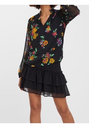 Kobieta nosząca czarną kwiecistą bluzkę z przezroczystymi długimi rękawami i czarną, warstwową mini spódniczkę z falbankami.