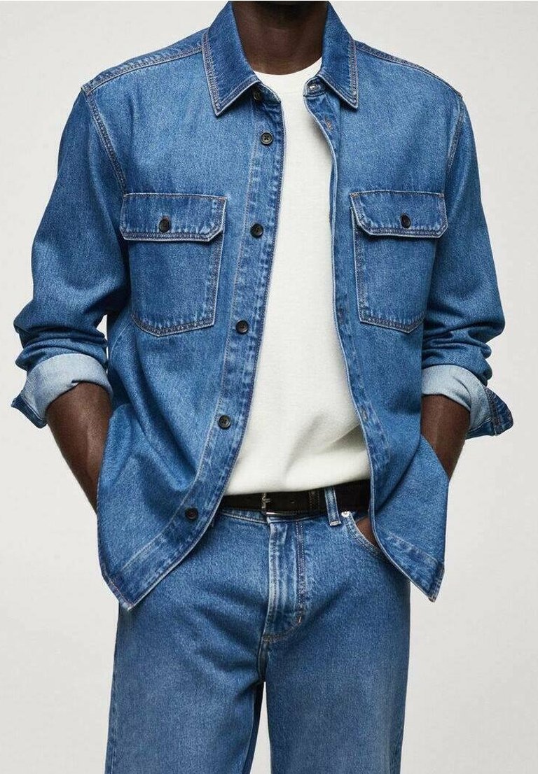 Homme portant une veste en denim bleue aux manches retroussées sur une chemise blanche, associée à un jean denim bleu et une ceinture noire, mains dans les poches.