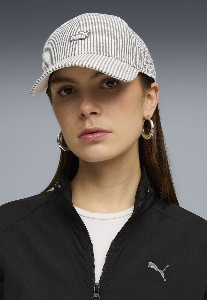 Femme portant une veste noire Puma zippée jusqu'à la poitrine et une casquette de baseball Puma blanche à rayures grises, ainsi que des boucles d'oreilles argentées en forme d'anneau.