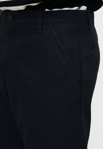 Κοντινή λήψη μαύρου ραμμένου παντελονιού με πλαϊνή τσέπη, θηλιές για ζώνη και κλείσιμο με κουμπί, φορεμένο με ριγέ μαύρο και άσπρο τοπ.