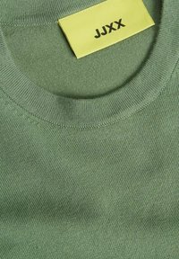 Maglione girocollo verde in cotone con collo a costine e un'etichetta gialla con lettering nero. Tessuto morbido con una finitura liscia.