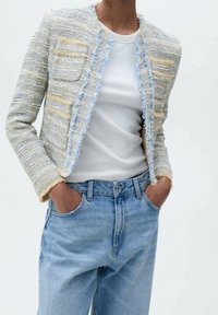 Veste en tweed texturée bleu clair et jaune avec des bords effilochés, deux poches avant, portée sur une chemise blanche et un jean bleu.