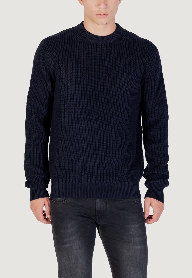 Maglione a coste navy con scollo rotondo, maniche lunghe e polsini aderenti, abbinato a jeans scuri, mettendo in risalto una superficie testurizzata.