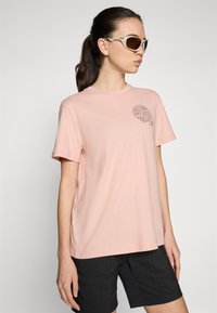 Lyserød bomuld t-shirt med korte ærmer, der har et sort grafisk design på venstre bryst. Båret med mørke shorts og solbriller.