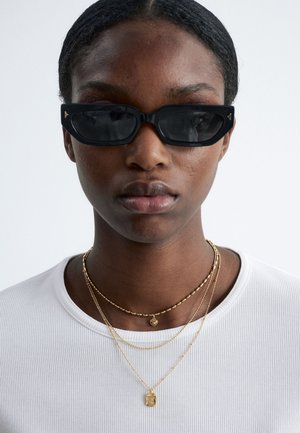 Sorte rektangulære solbriller med tonede glas, båret over en hvid ribbet trøje, ledsaget af flere gyldne lagdelte halskæder.