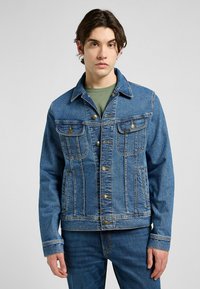 Chaqueta de mezclilla en azul medio, con cierre de botones en la parte frontal, dos bolsillos en el pecho y bolsillos laterales, con un cuello clásico y detalles de costura visibles.