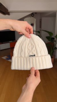 Fehér kötött sapka bordázott szegéllyel, fonott mintázattal és elöl fekete "Coach" logóval. Téli viseletre alkalmas.