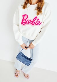 Vit sweatshirt med glittrande rosa "Barbie"-logga, ljusblå denimjeans, blå denimaxelväska och ljusrosa sandaler med remmar.