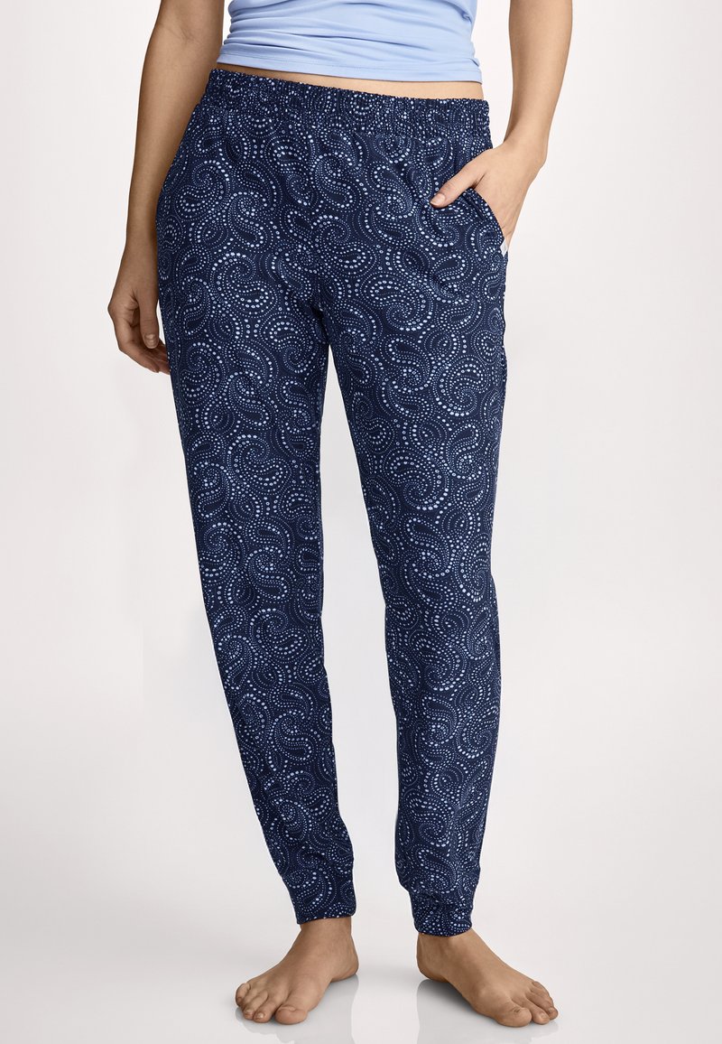 Vrouw draagt marineblauwe joggerbroek met lichtblauwe, gestippelde krulpatronen en elastische tailleband, staat blootsvoets met één hand in de zak.